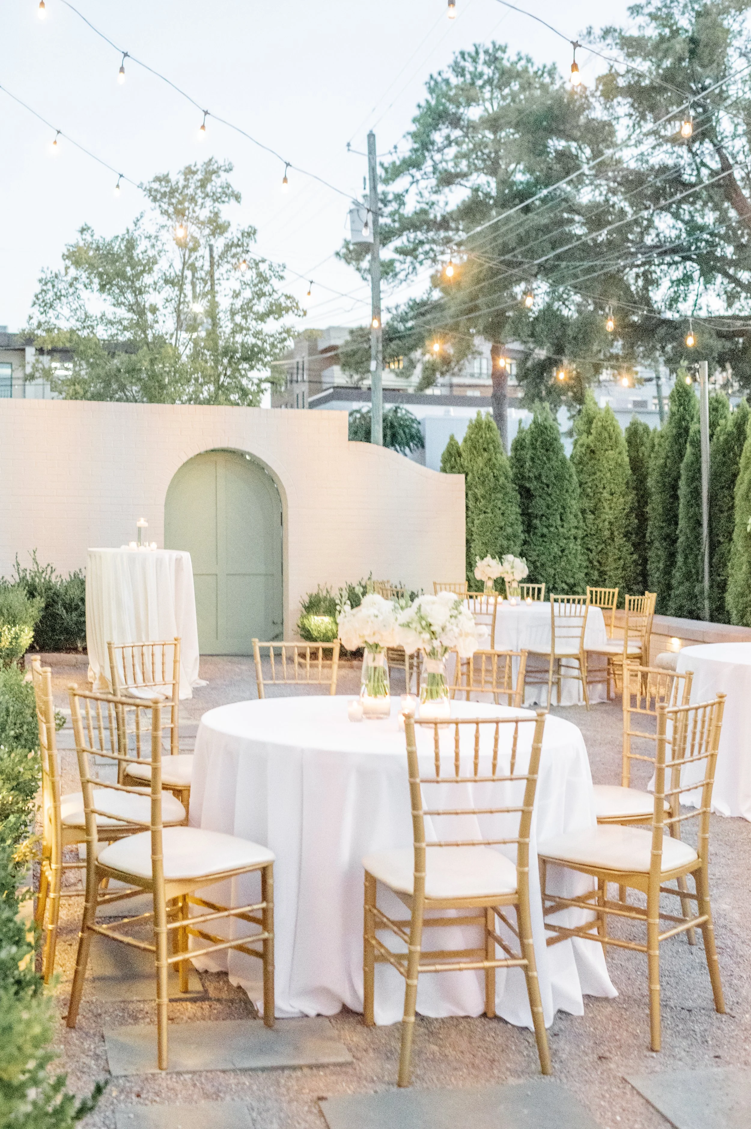 Outdoor event space with round tables covered in white tablecloths, decorated with white floral centerpieces in glass vases, surrounded by gold chairs, under string lights, with a white brick wall and greenery in the background.