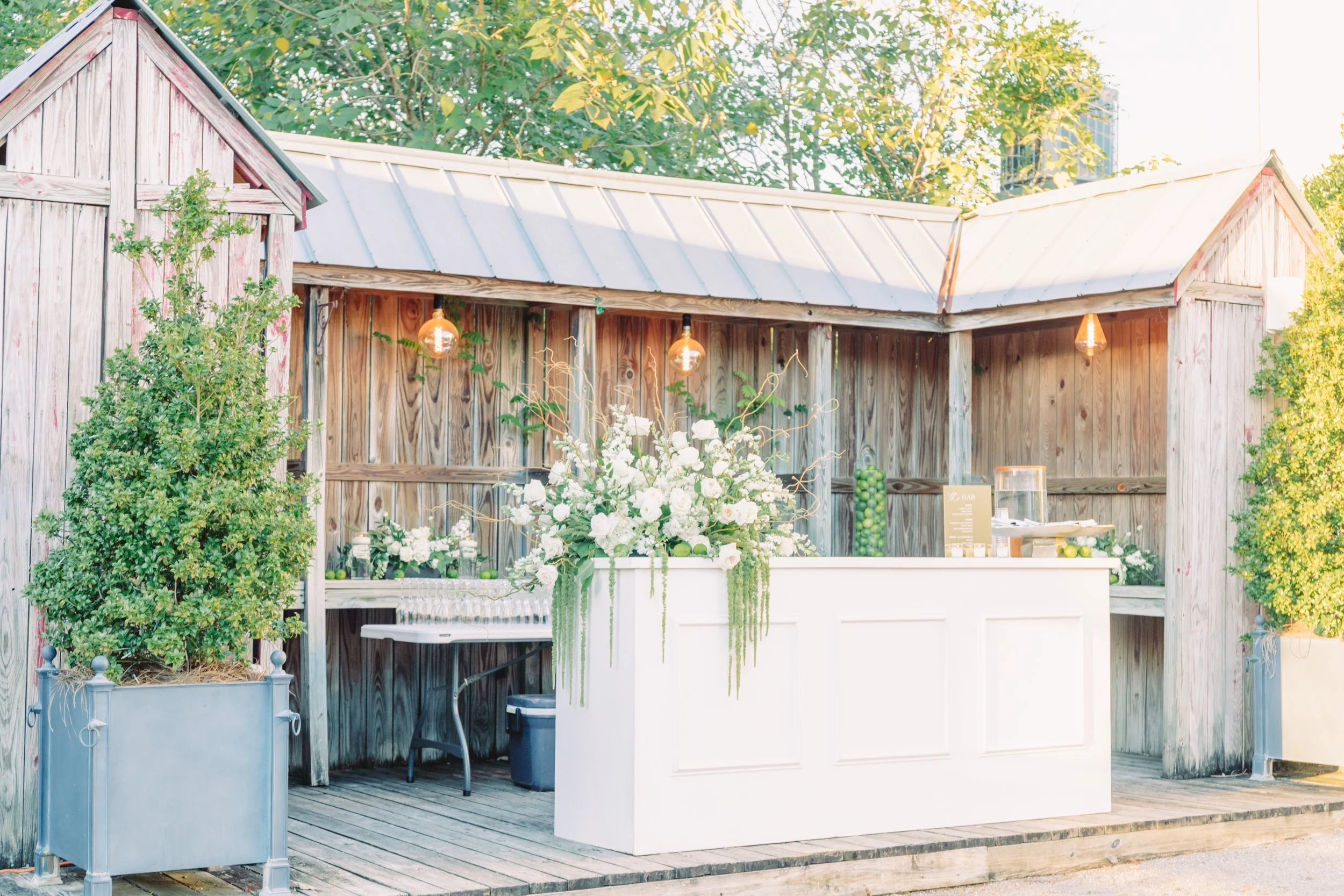 Outdoor bar area with a wooden backdrop, decorated with string lights, large floral arrangement, and greenery, set on a wooden platform.