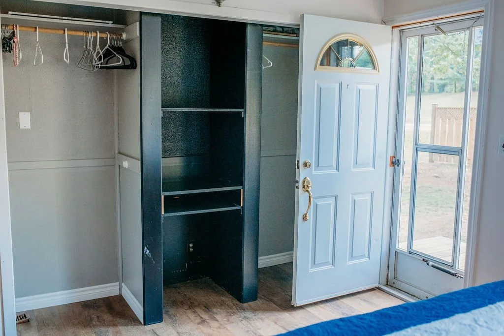 Empty closet with hanging rod and shelves, next to a white door leading outside, with a view of a backyard.