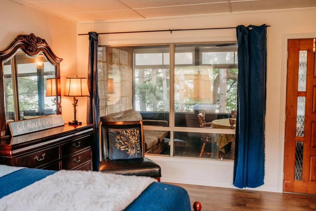 A cozy bedroom with a wooden dresser and mirror, bedside lamp, and a black leather chair with a decorative pillow. Large glass sliding doors with blue curtains lead to a screened porch with a round table and chairs, overlooking a wooded area.