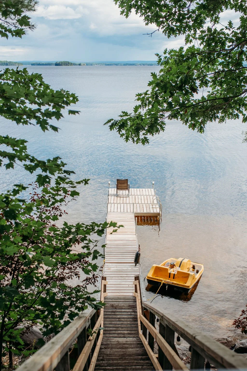 A wooden staircase leading down to a dock on a lake, with a yellow paddle boat tied to the dock, surrounded by green leaves, and distant trees on the horizon.