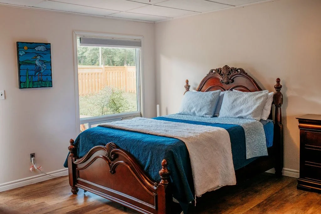 A bedroom with a wooden bed frame, blue and white bed linens, a window showing a wooden fence and greenery outside, a colorful painting on the wall, and a small dark wooden nightstand.