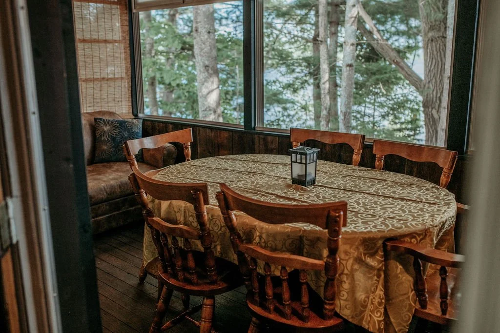 A cozy dining nook with a round table covered in a patterned tablecloth, surrounded by six wooden chairs, and a window view of dense green trees outside.