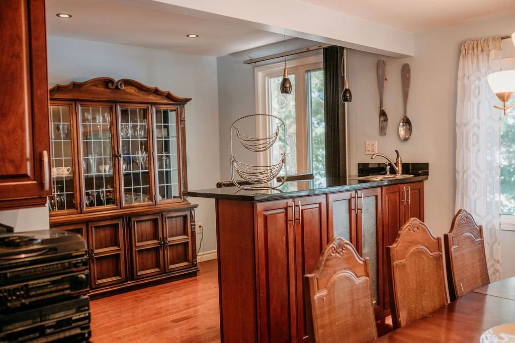 A cozy dining area with a wooden dining table and chairs, a wooden china cabinet, a kitchen island with a sink, hanging utensils on the wall, and large windows with light curtains letting in natural light.