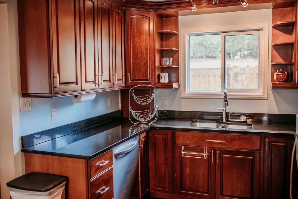 Kitchen with wooden cabinets, black granite countertops, a stainless steel sink, a window over the sink, and open shelves on both sides of the window.