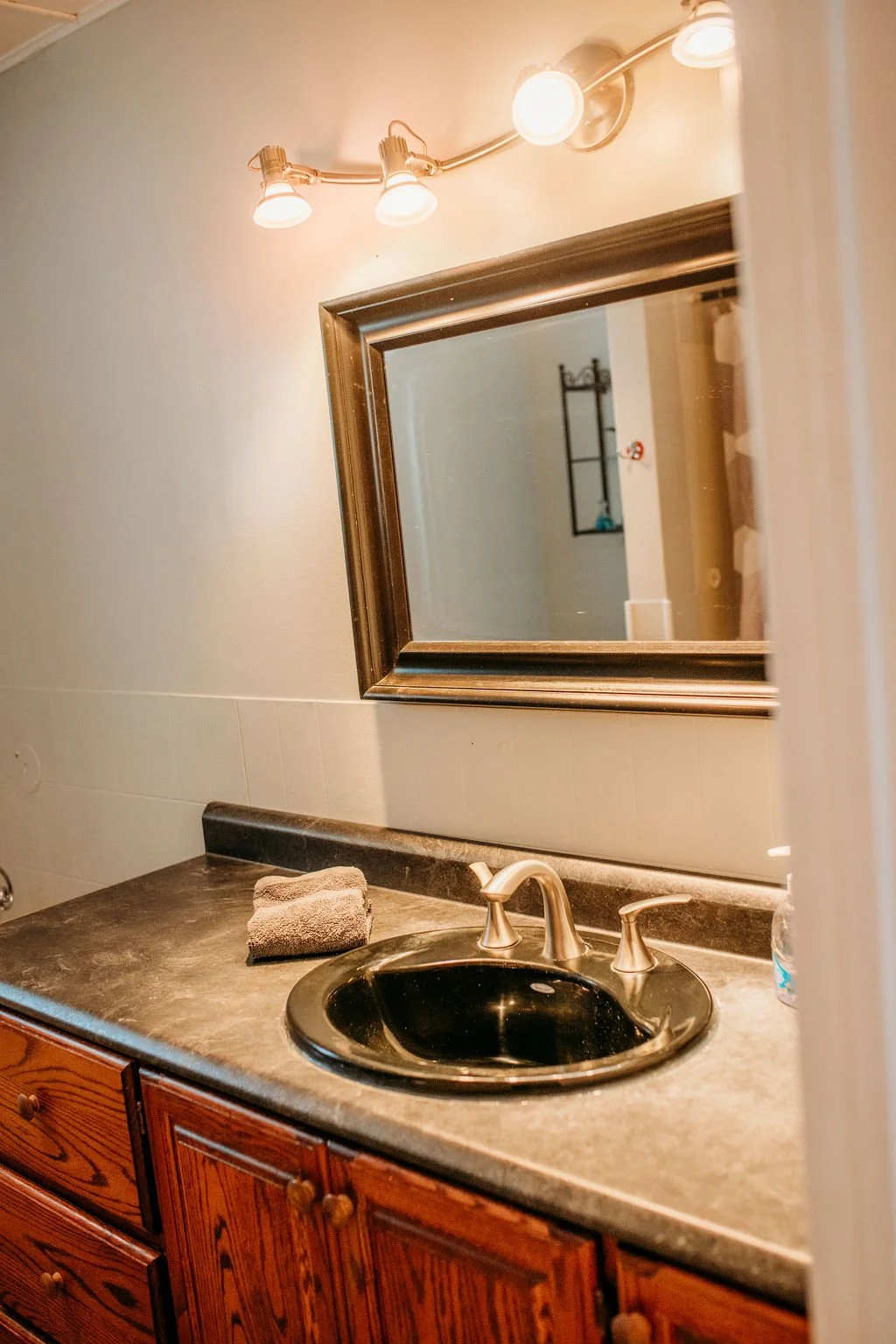 A bathroom vanity with a black sink, a faucet, and a mirror above it. There is a folded towel on the countertop and a light fixture with three bulbs above the mirror. Wooden cabinets are below the countertop.
