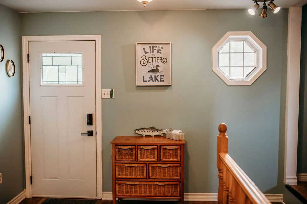 Entrance area of a home with a white door, a light blue wall, a small wooden chest with a fish decoration and a tissue box, a wall-mounted sign reading "Life is better at the lake," a small octagon window, and track lighting on the ceiling.