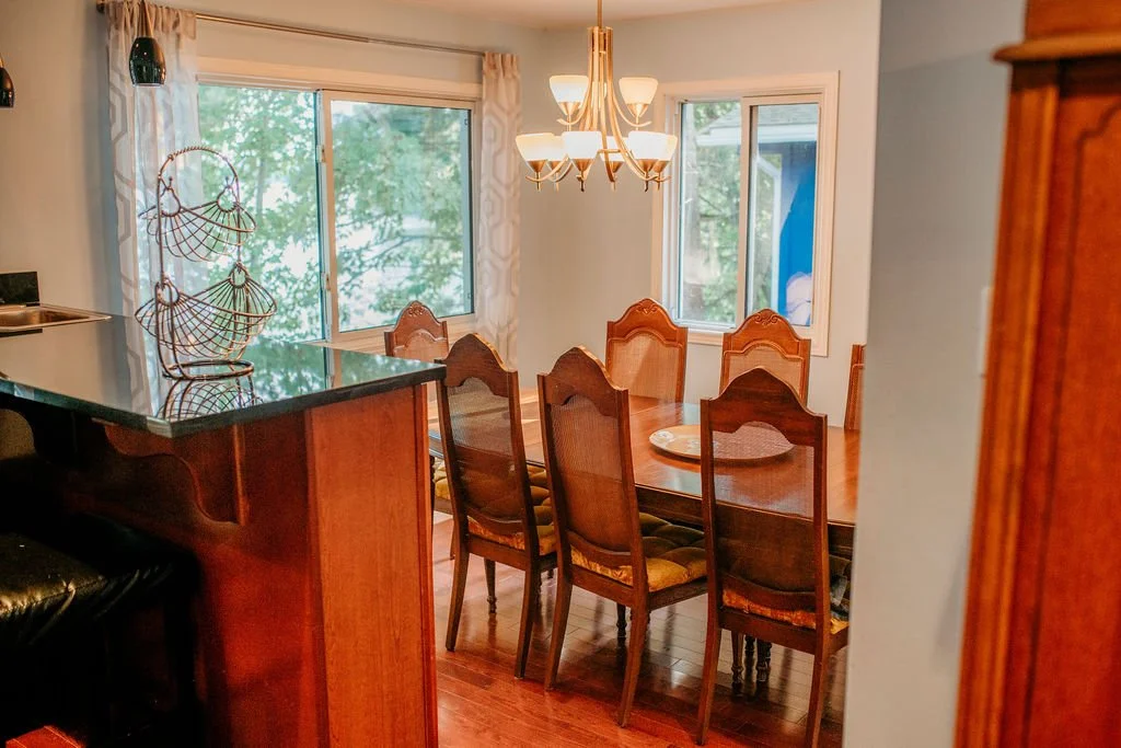 A dining area with a wooden table and six wooden chairs with patterned cushions, a chandelier hanging above, two windows showing outdoor greenery, and curtains with a light pattern.