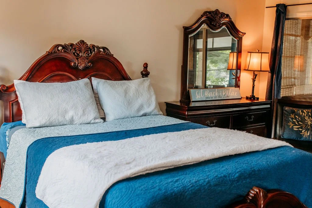 A bedroom with a wooden bed frame, white pillows, and a blue and white quilt, a dark wood dresser with a mirror, a lamp, and a decorative sign that reads 'Sit back & relax, you're all set to shine.'