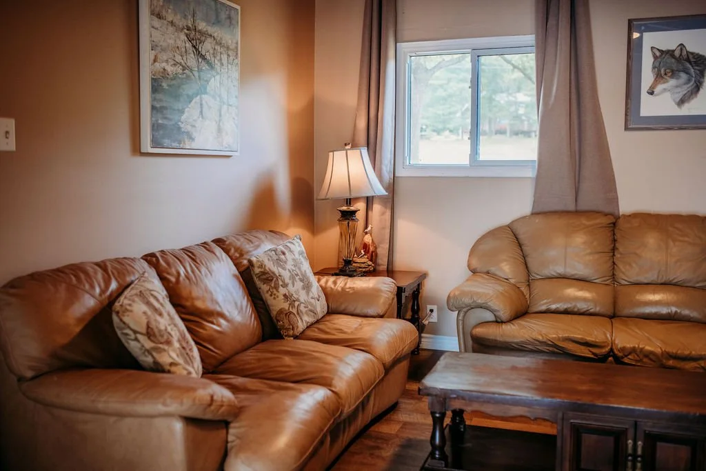 Living room with leather sofas, a wooden coffee table, a window with brown curtains, and wall art, including a landscape and a wolf portrait.