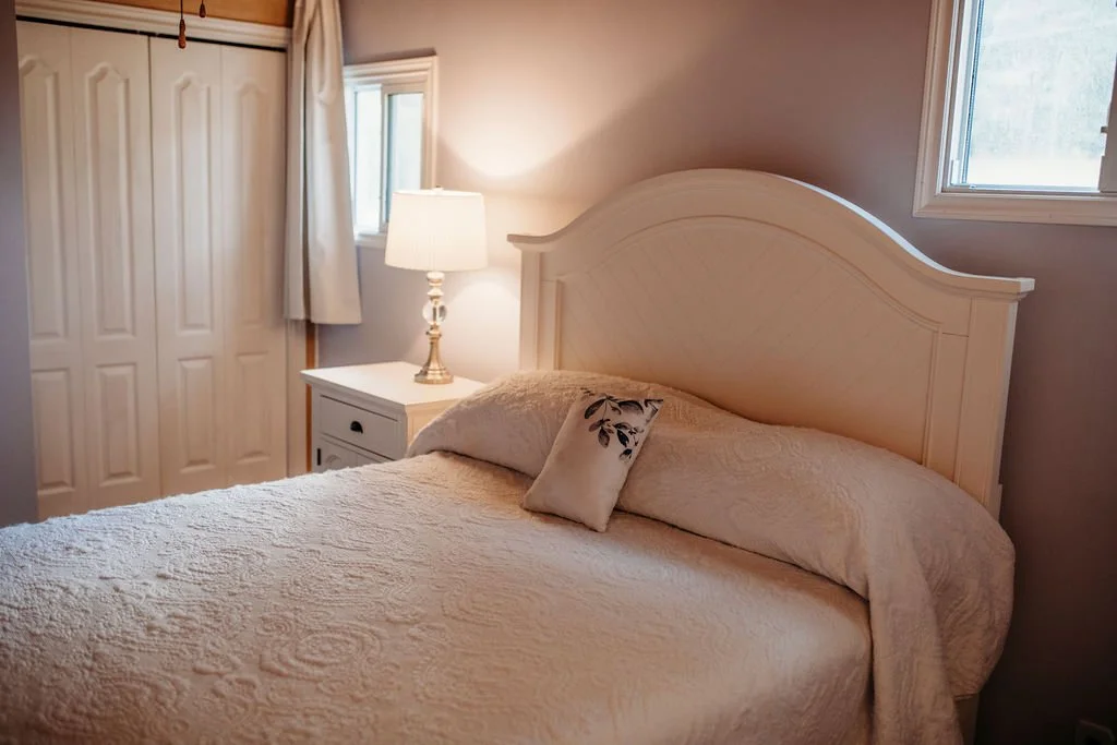 A cozy bedroom with a large bed, cream-colored quilt, decorative pillow, white headboard, white nightstand with a lamp, and windows with cream curtains.