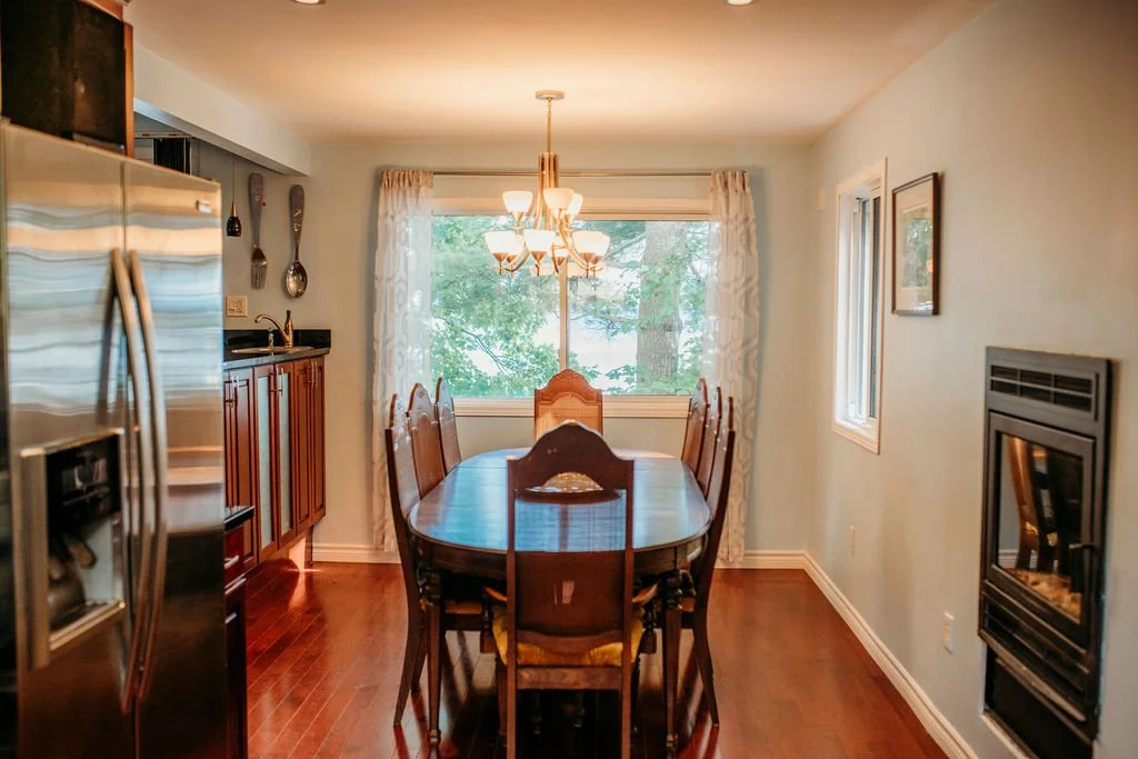 Dining room with a wooden table and eight chairs, a chandelier hanging above, a large window with curtains showing trees outside, a side sideboard with utensils, and a stainless steel refrigerator on the left.