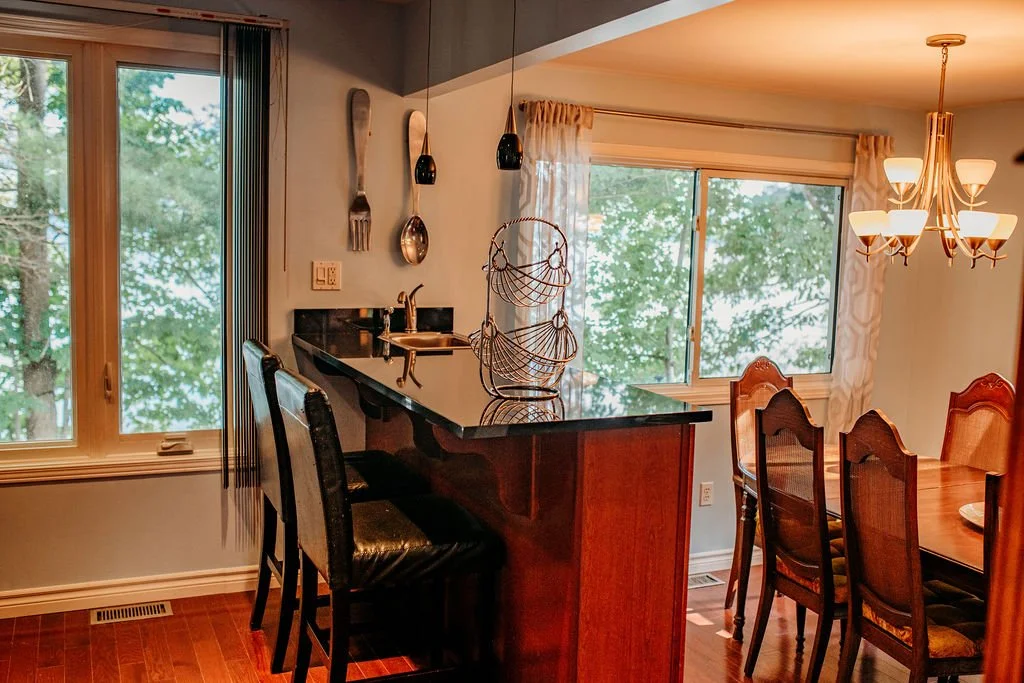 A kitchen island with black countertop and wooden base, with a small sink and a wire basket on top. Two black chairs are placed by the island, and there are large windows with trees visible outside. To the right, a dining area with a wooden table and