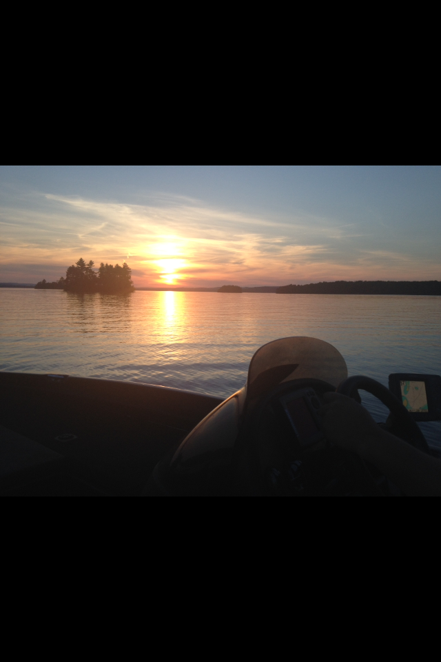 Sunset over a body of water with a silhouetted island and trees, view from inside a boat with a steering wheel and GPS device visible.