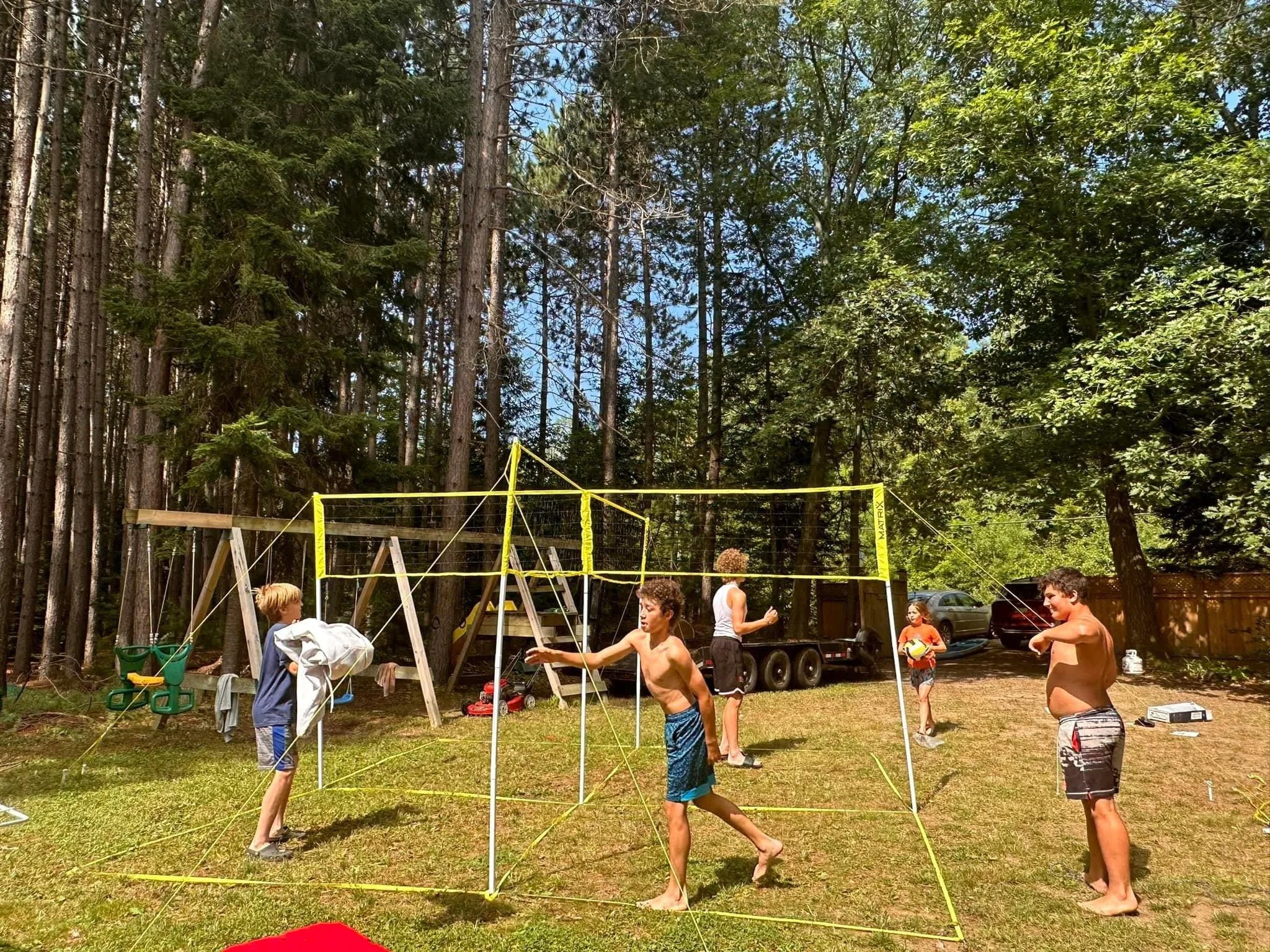 Children playing in a backyard volleyball game on a sunny day, surrounded by trees, with cars parked in the background.
