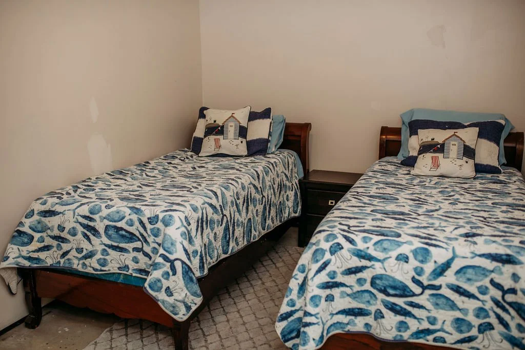 Two twin beds with beach-themed bedding and coastal pillows in a small bedroom with plain walls.