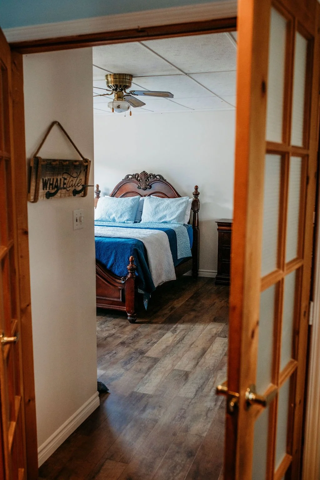 A bedroom viewed through a doorway, featuring a wooden bed with blue and white bedding, a ceiling fan, a dark wooden dresser, and wood flooring.