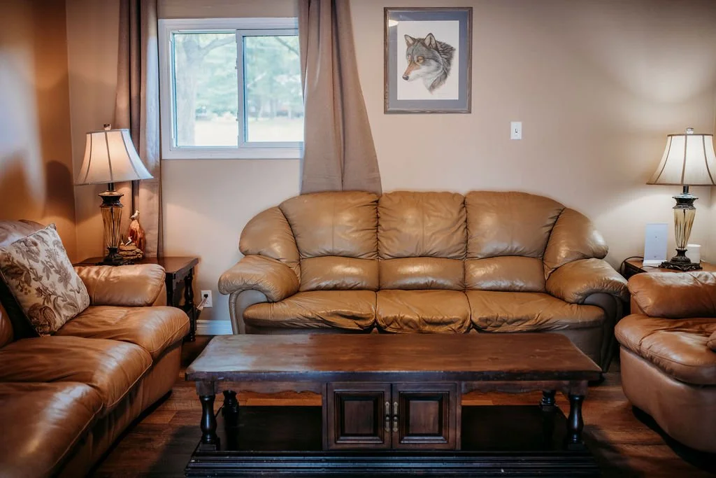 Living room with brown leather sofa, two matching armchairs, two lamps, a small wooden side table, a framed wolf portrait on the wall, a window with beige curtains, and a wooden coffee table.