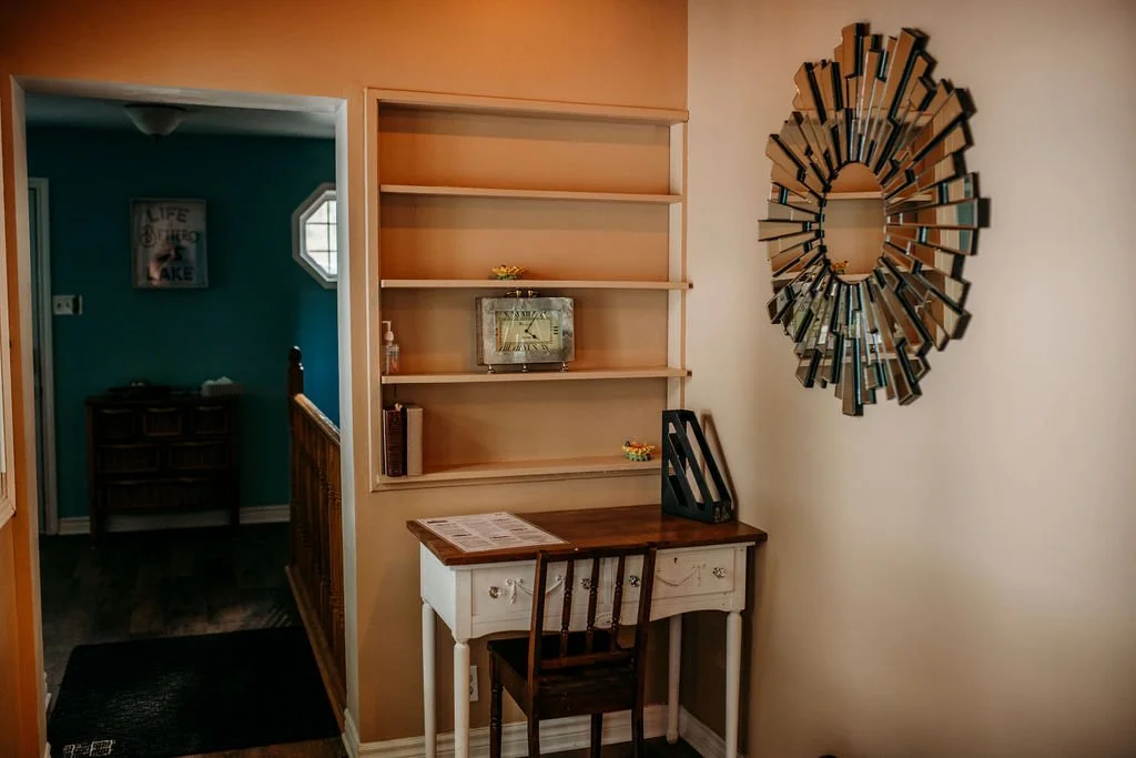 Interior view of a room with a decorative mirror, a desk, and empty shelves. An open doorway shows another room with a small side table and framed photo.