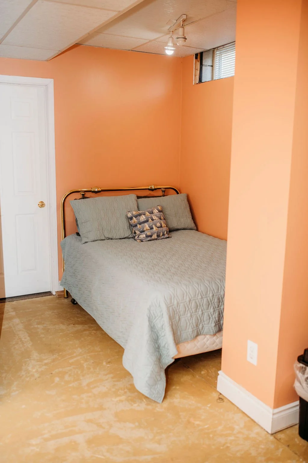 A bedroom with peach-colored walls, a metal bed frame, a light gray quilt, and three pillows, one with a pattern of sailboats. There is a white door on the left, a window with blinds above the bed, and a small window near the ceiling. The floor is unfinished wood or concrete.