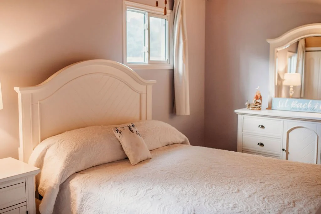 A cozy bedroom with a white bed, beige bedding, and a single decorative pillow. There's a window with beige curtains, a white dresser with a mirror, and a blue sign that reads 'Still Beach' on top of the dresser. The room has warm lighting.