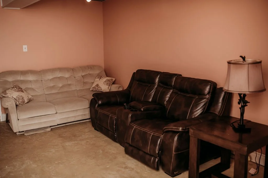 Living room corner with beige tufted sofa, dark brown leather recliner, wooden side table, and table lamp with beige shade, wall painted in light pink.
