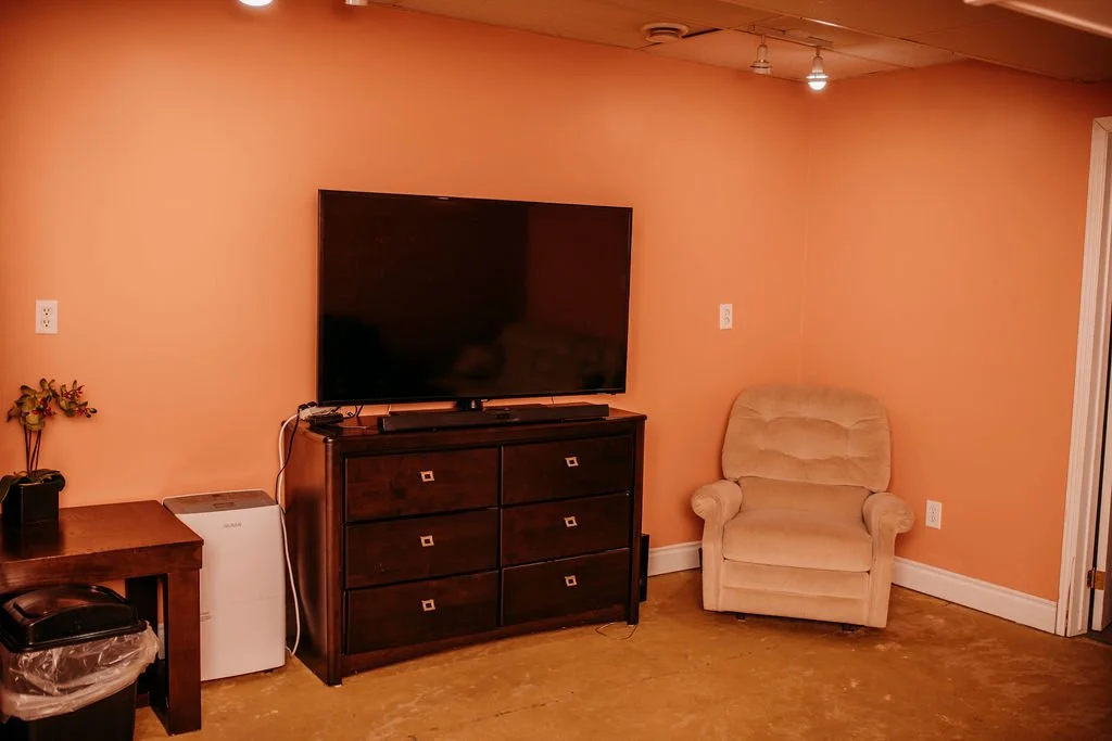 Living room with a large flat-screen TV on a wooden dresser, a beige armchair, a small wooden table with flowers, a mini air purifier, and a doorway to the right, all against peach-colored walls.