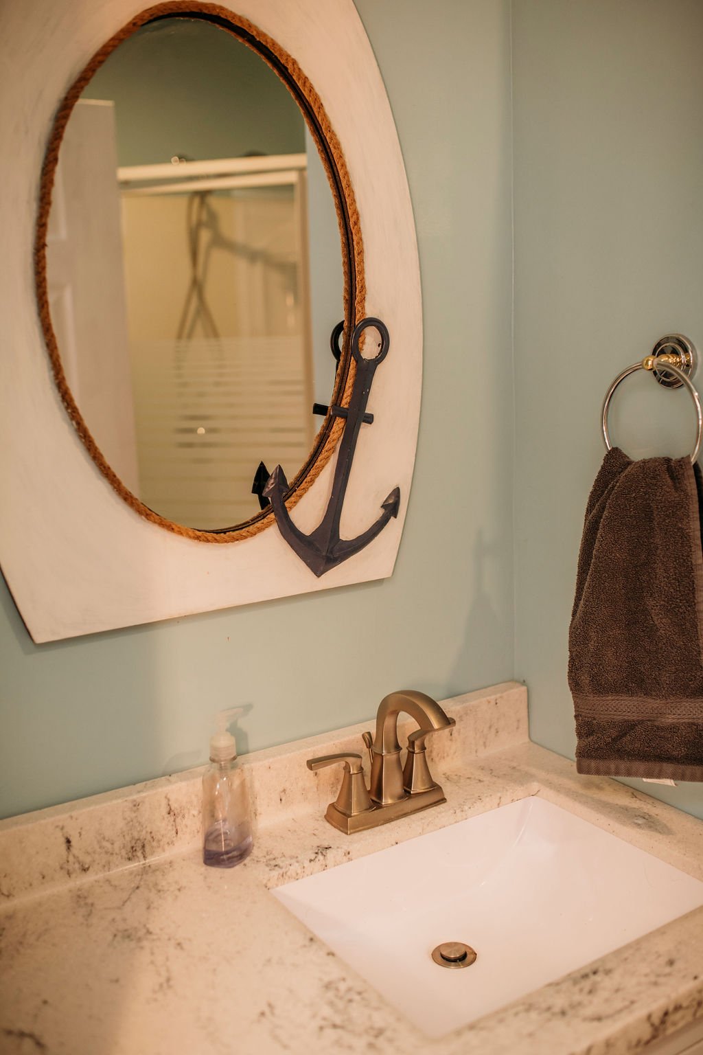 Bathroom with oval mirror featuring a black anchor decoration, beige granite countertop, white sink, bronze faucet, brown towel hanging on a towel ring, and a soap dispenser.