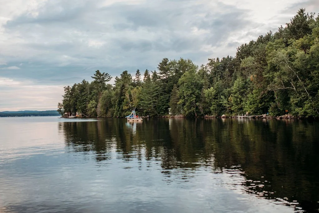 Calm lake surrounded by dense green trees with a small boat floating near the shoreline under a cloudy sky.