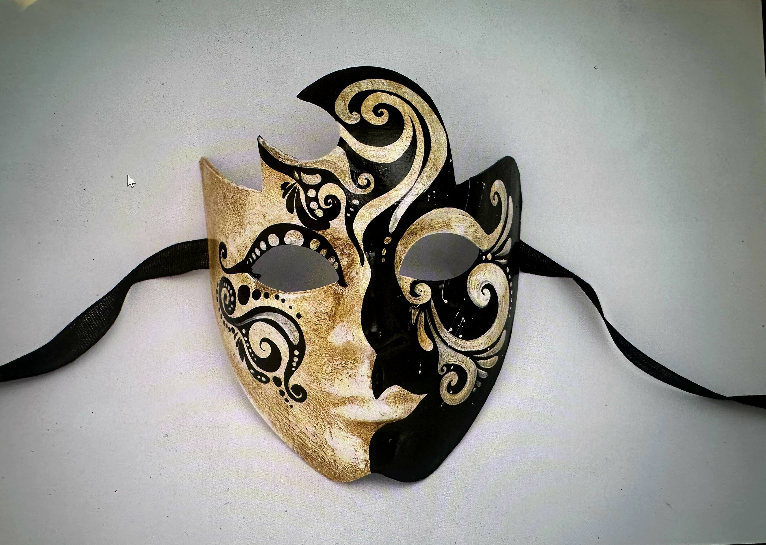 A decorative half-face mask with intricate black swirling patterns and beige face part, mounted when displayed on a plain white surface.