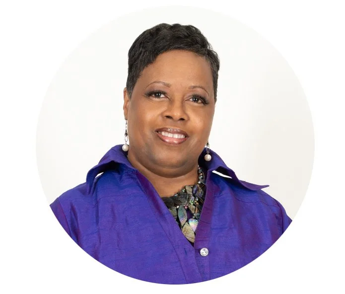 A portrait of a woman with short dark hair, wearing a purple shirt, pearl earrings, and a necklace, smiling against a white background.