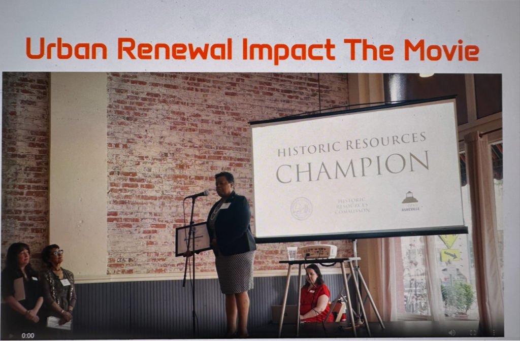 A woman speaking at a public event with a microphone, with three women sitting to her left and a large screen behind her displaying 'HISTORIC RESOURCES CHAMPION.' The event is related to urban renewal and historic resources.