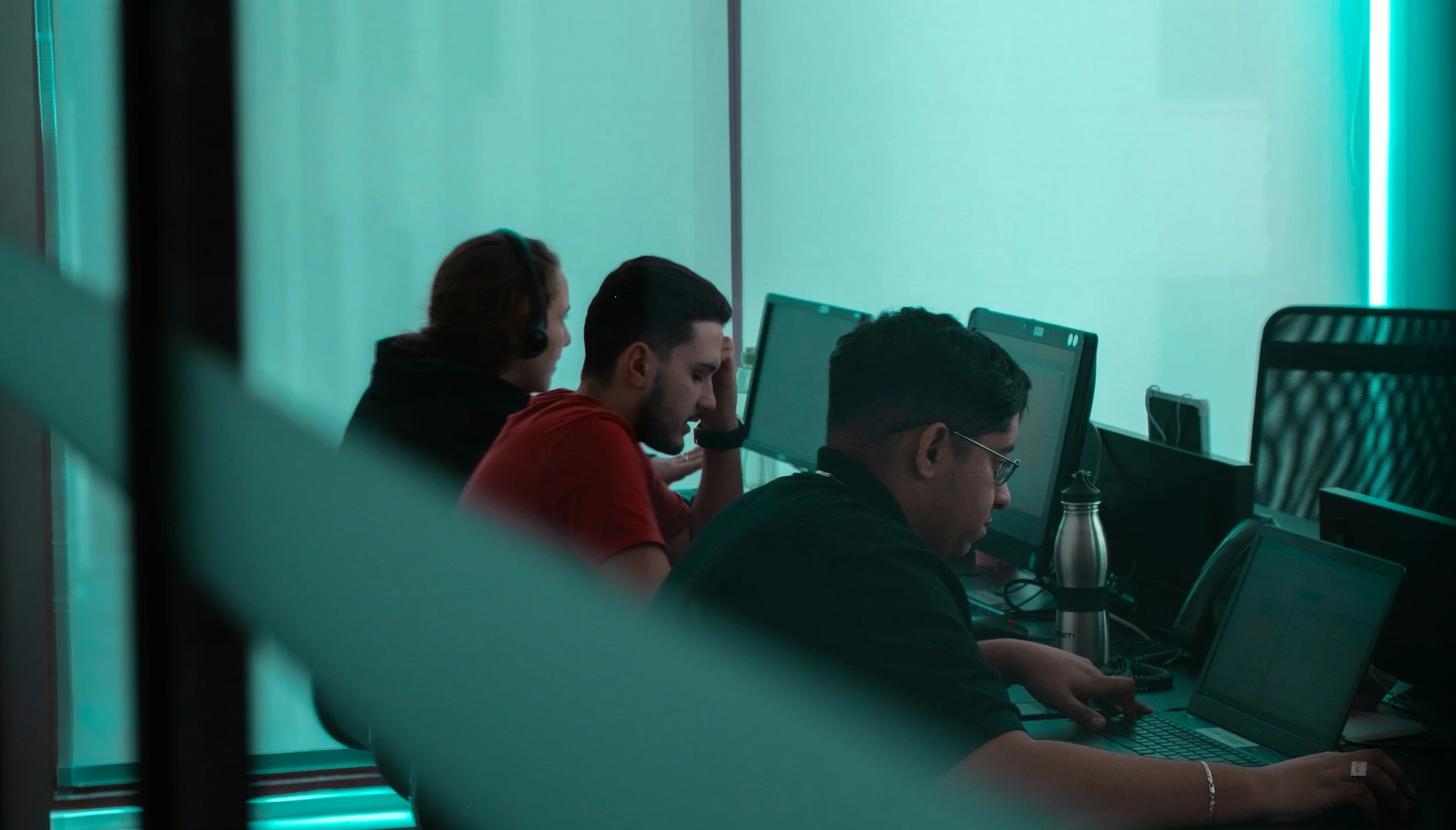 Three people working at desks with computer monitors in a modern office with teal lighting.
