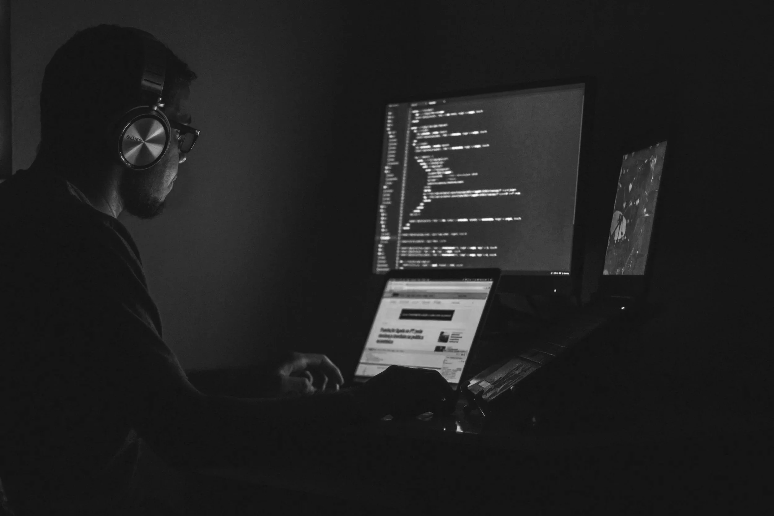 Person wearing headphones working on a laptop with dual monitors in a dark room.