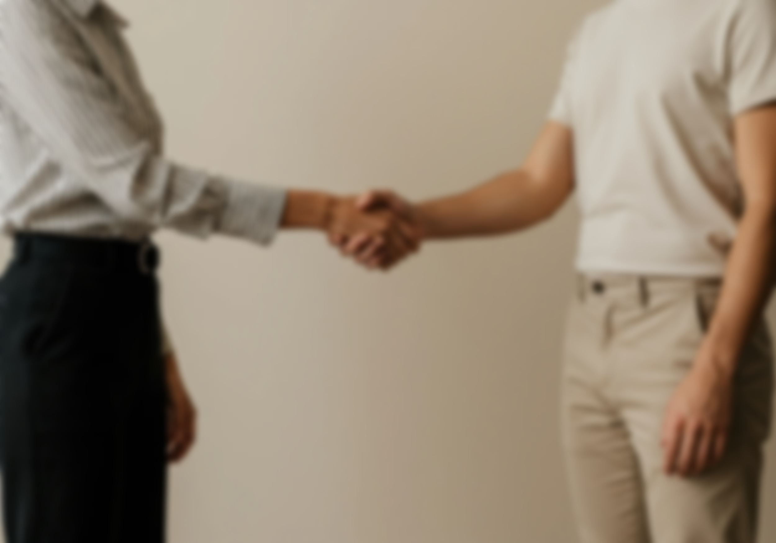 Two people are shaking hands in a professional setting. One person is wearing a striped shirt and dark pants, the other is wearing a white t-shirt and beige pants. The background is plain and neutral.