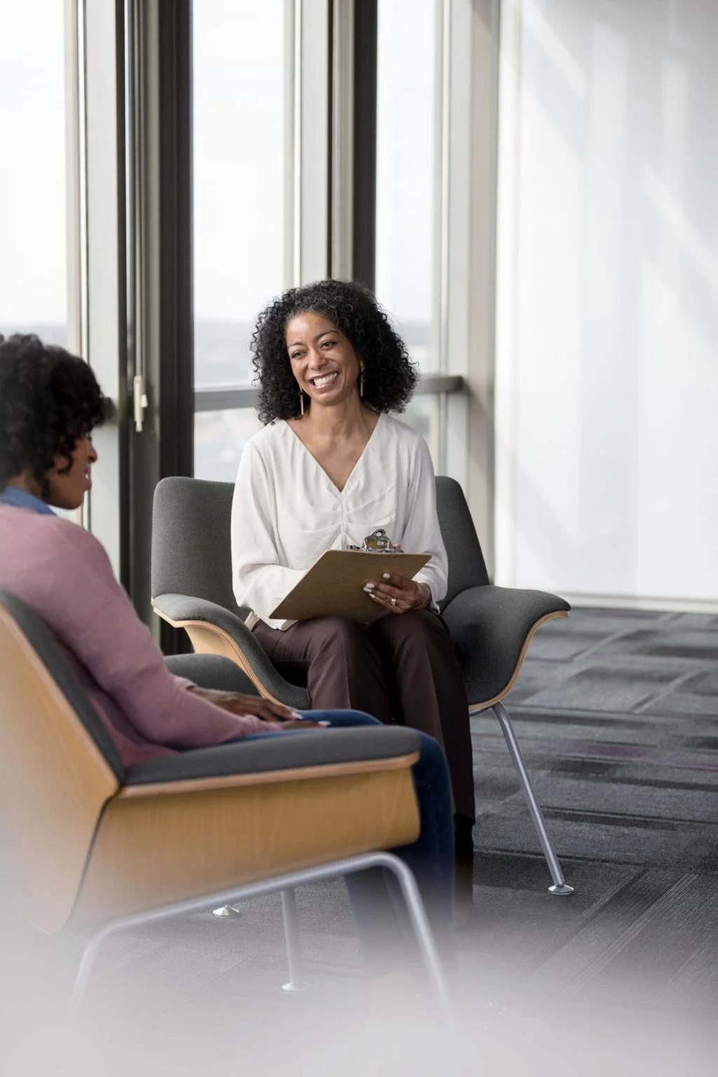 Two women sitting in a modern office, one smiling and holding a clipboard, engaged in conversation near large windows.