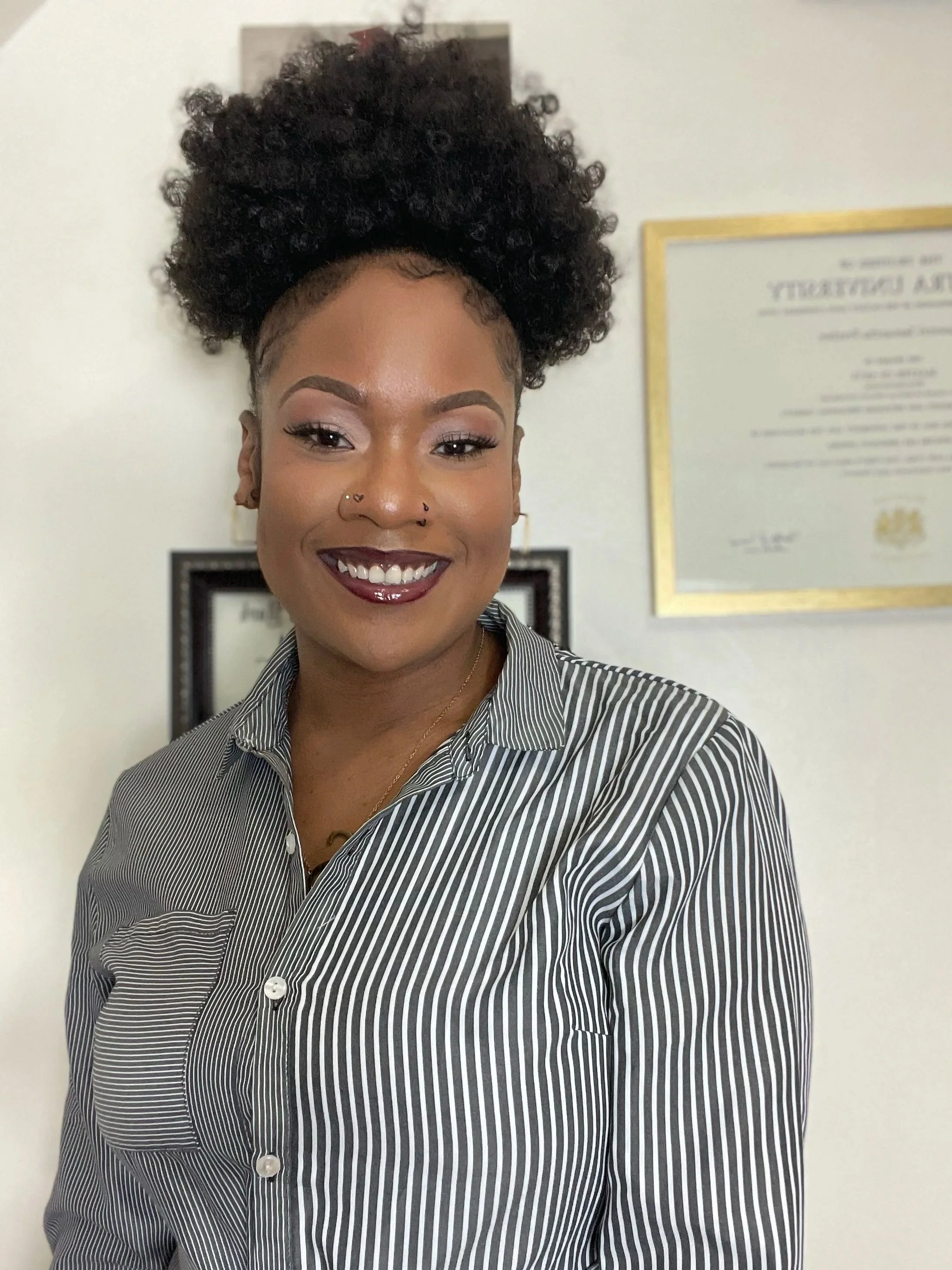 A Black woman with curly black hair styled in an updo, wearing a black and white pinstripe button-up shirt, smiling in front of a wall with framed license and degree documents and a gold border.