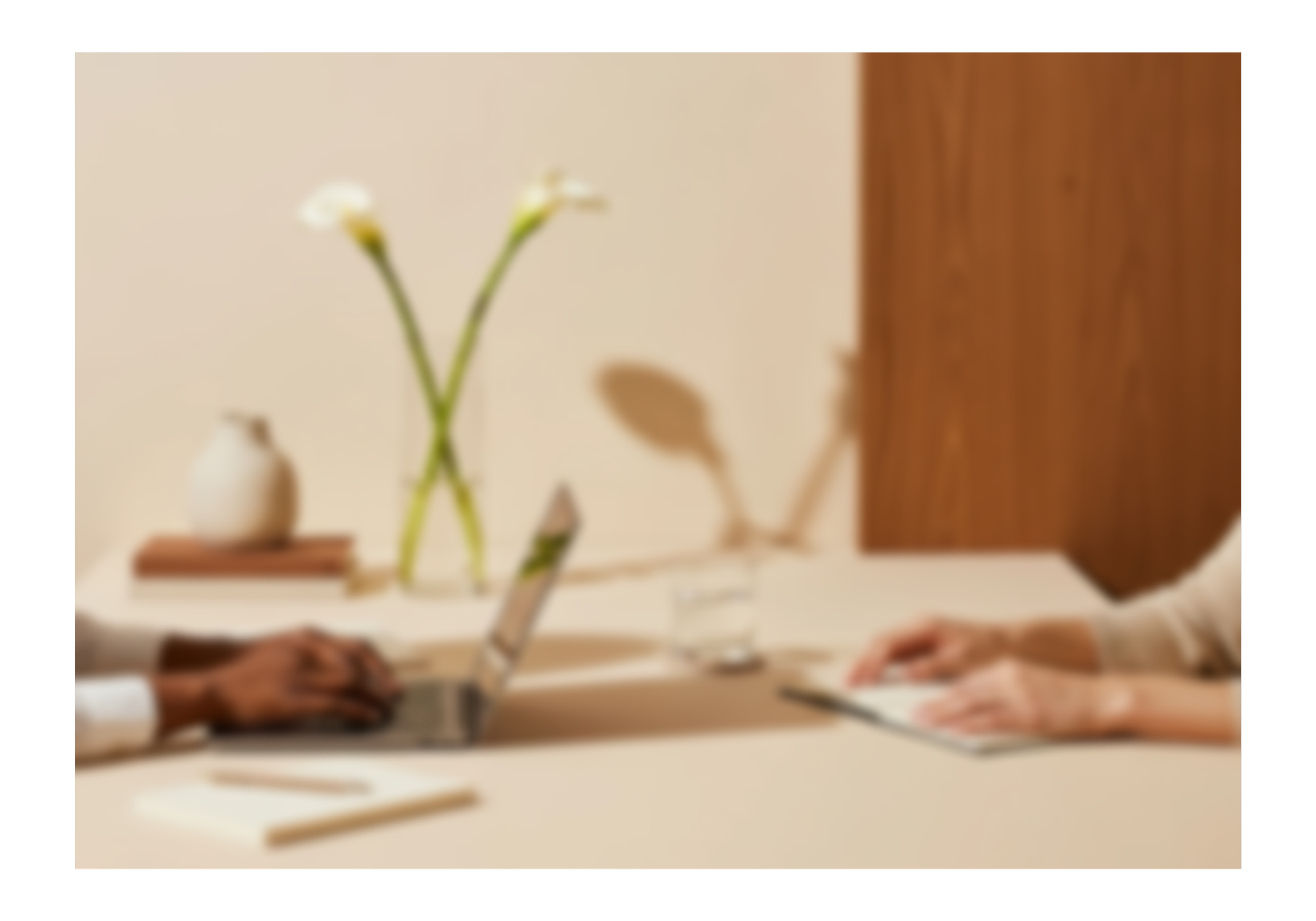 Two people sitting across from each other at a table, working on laptops. On the table, there are notebooks, a pen, a glass of water, and a small vase with two calla lily flowers. The background includes a beige wall with a wooden panel.