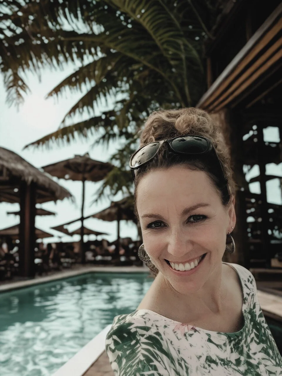 A woman smiling by a pool at a tropical resort, wearing sunglasses on her head and earrings, with palm trees and thatched umbrellas in the background.
