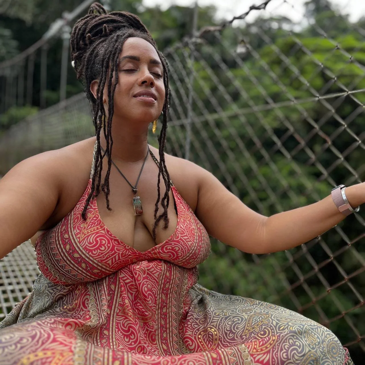 A woman with dreadlocks, dressed in a patterned dress, sitting on a suspension bridge with her eyes closed and a peaceful expression. A happy Luna & Soul client