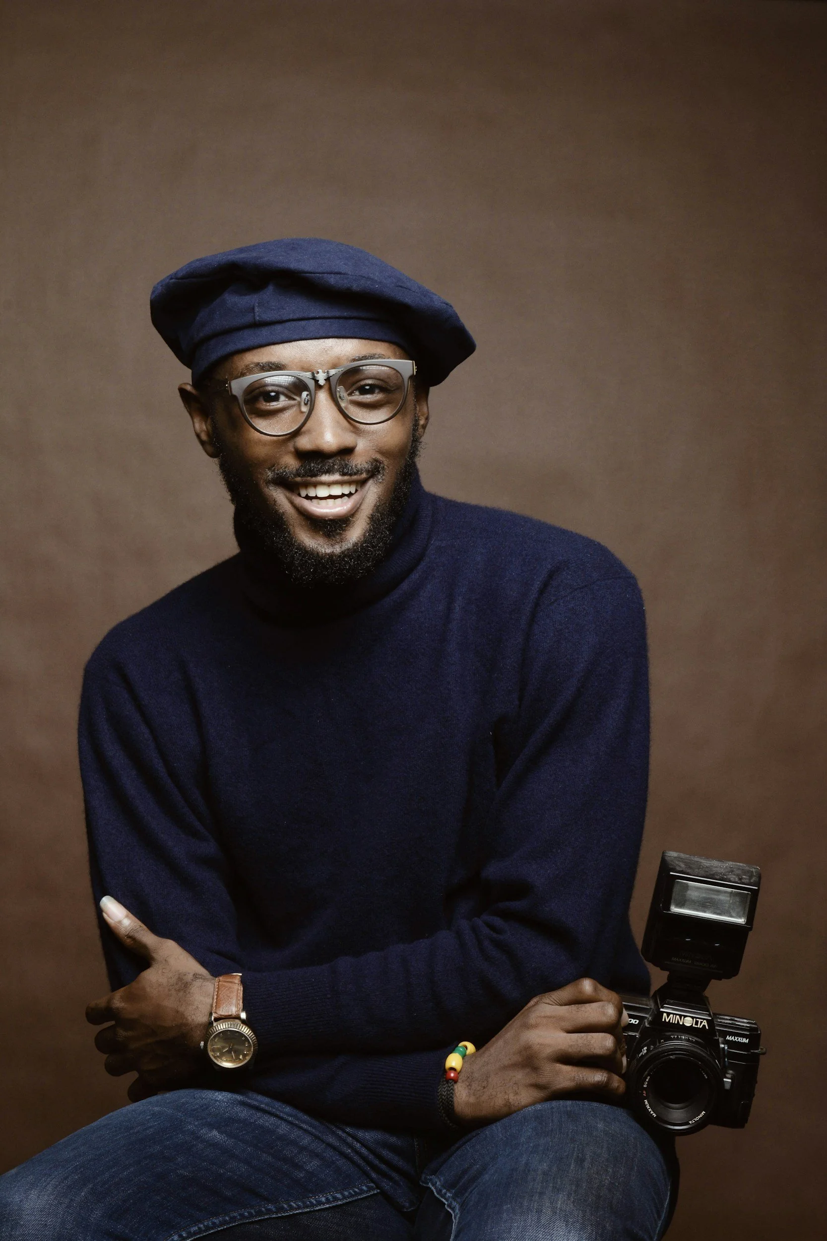Portrait of a smiling man wearing glasses, a beret, a navy blue sweater, and jeans, holding a vintage camera with a flash, against a brown background. A happy Luna & Soul client. 