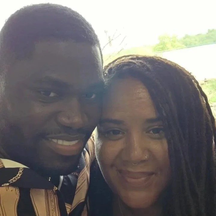 A close-up photo of a smiling Black man and woman with dreadlocks, taking a selfie together outdoors.