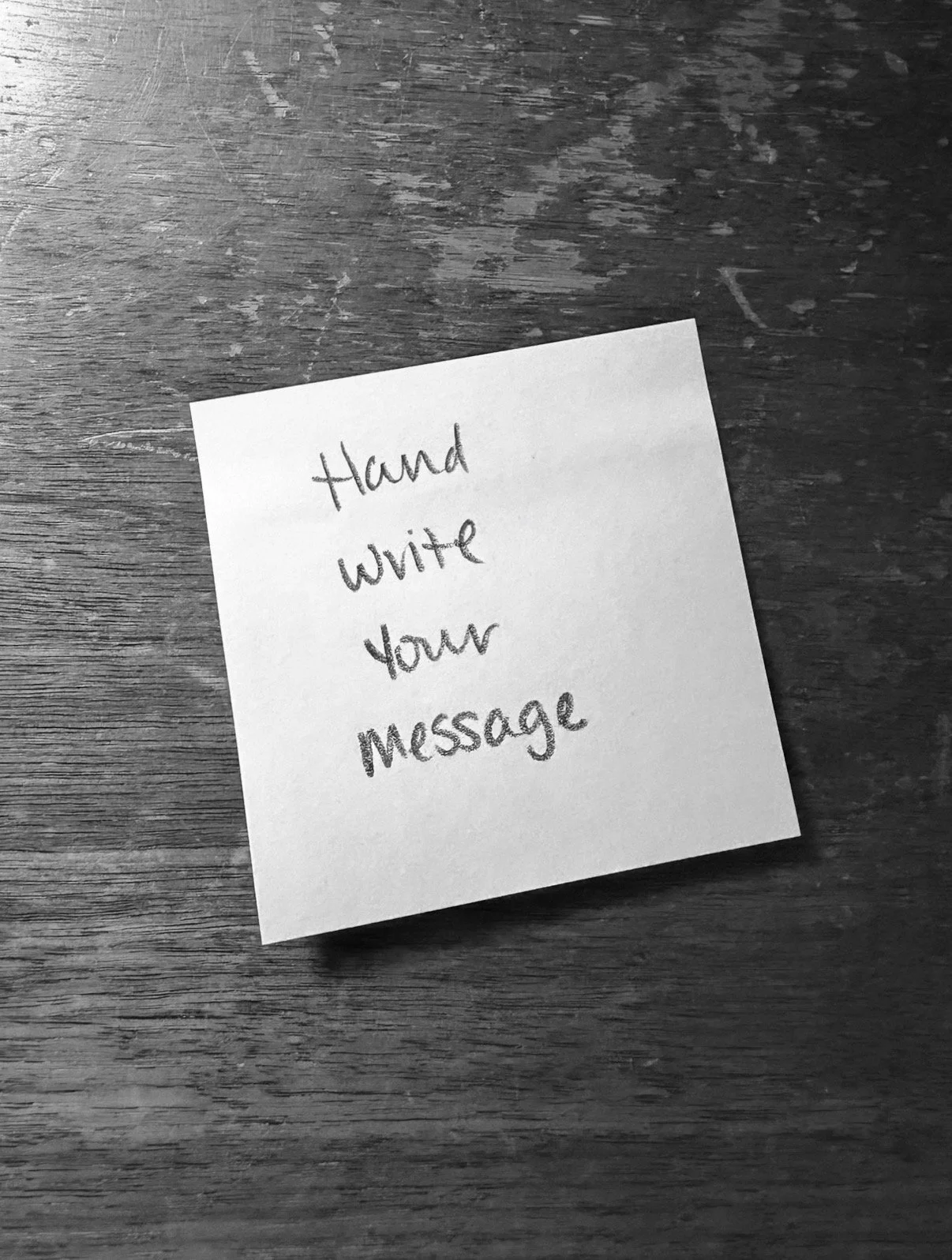"Hand write your message" written on a sticky note on a wood desktop photo in black and white