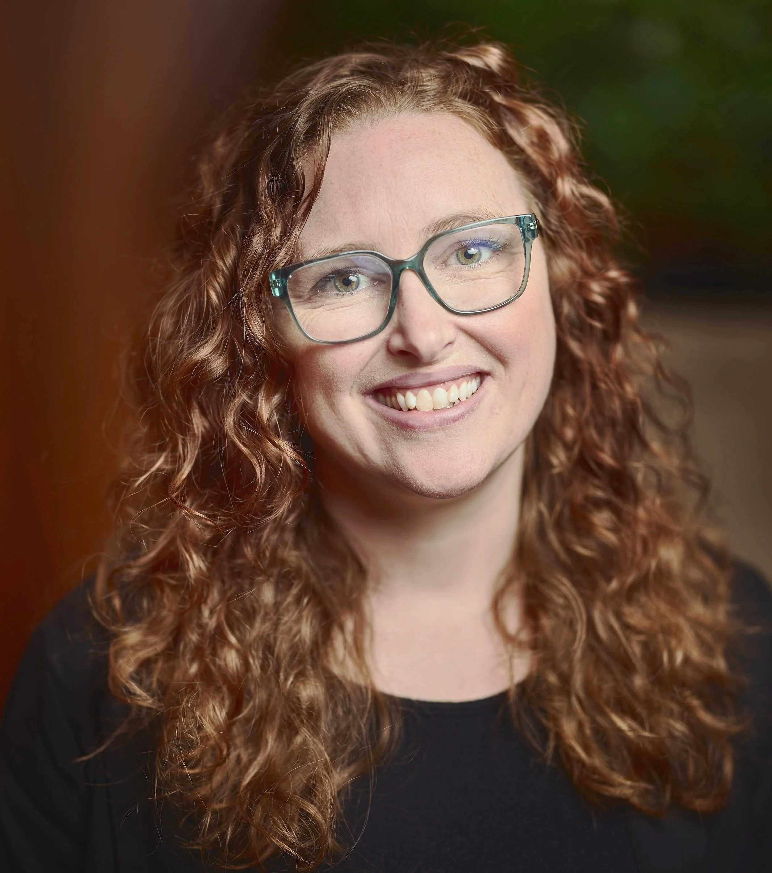 Headshot of a red, curly haired woman wearing a grey suit jacket, black top, and turquoise rimmed glasses