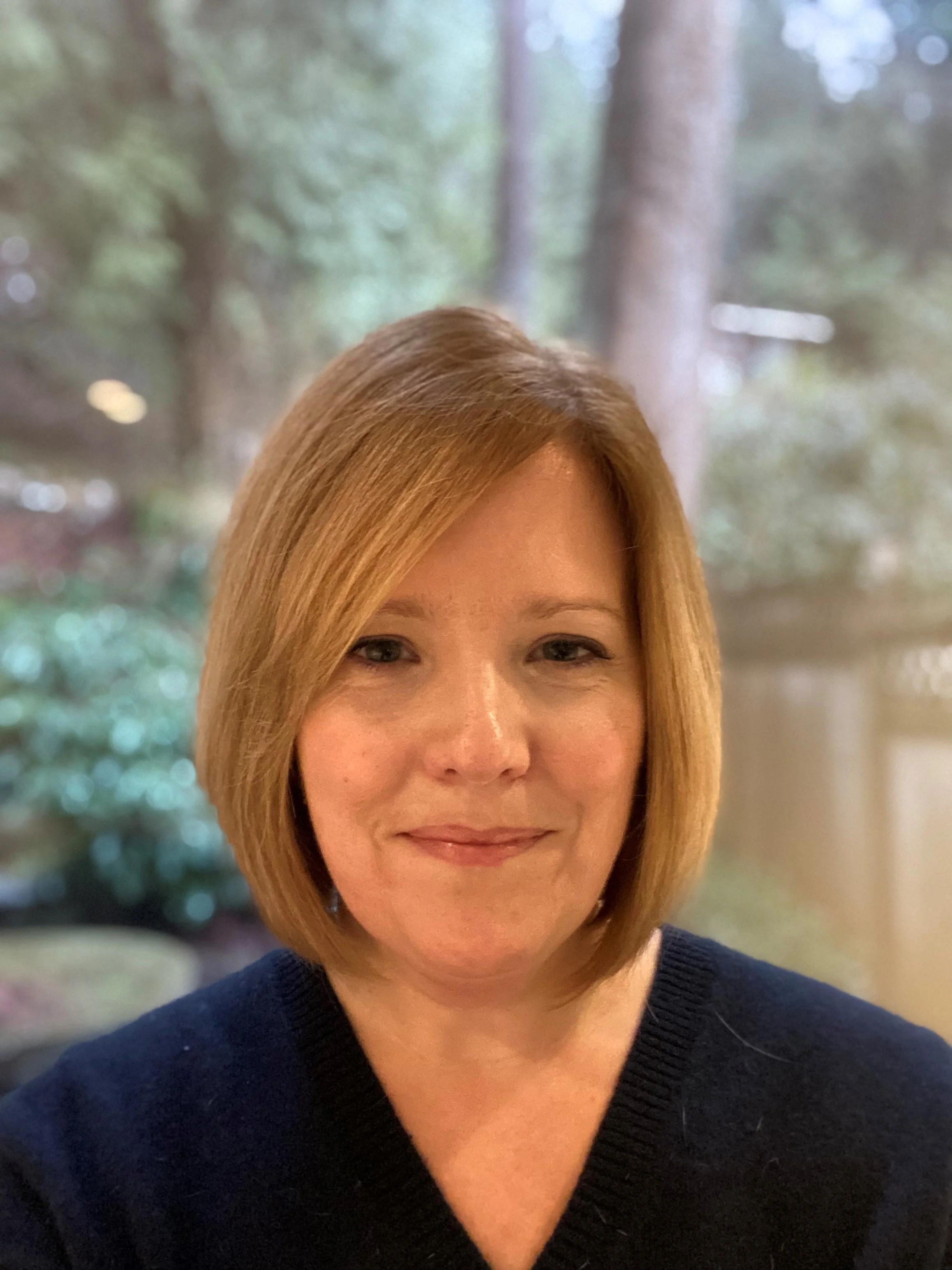 Casual headshot of a white woman, standing in front of a tree and fence, wearing a blue shirt, smiling at the camera, she has blond hair