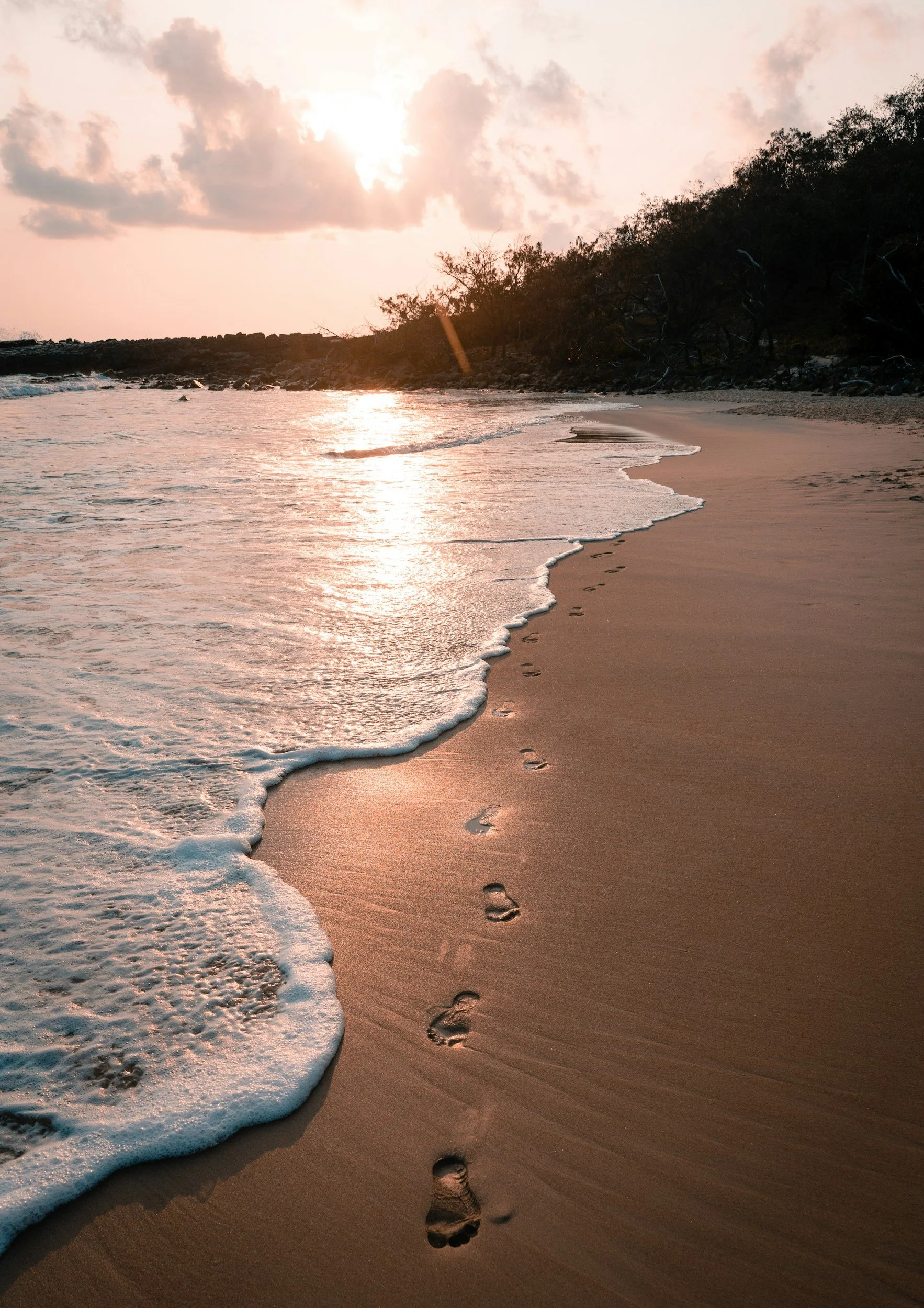 Footprints in the sand on a beach at sunset with gentle waves and a cloudy sky.