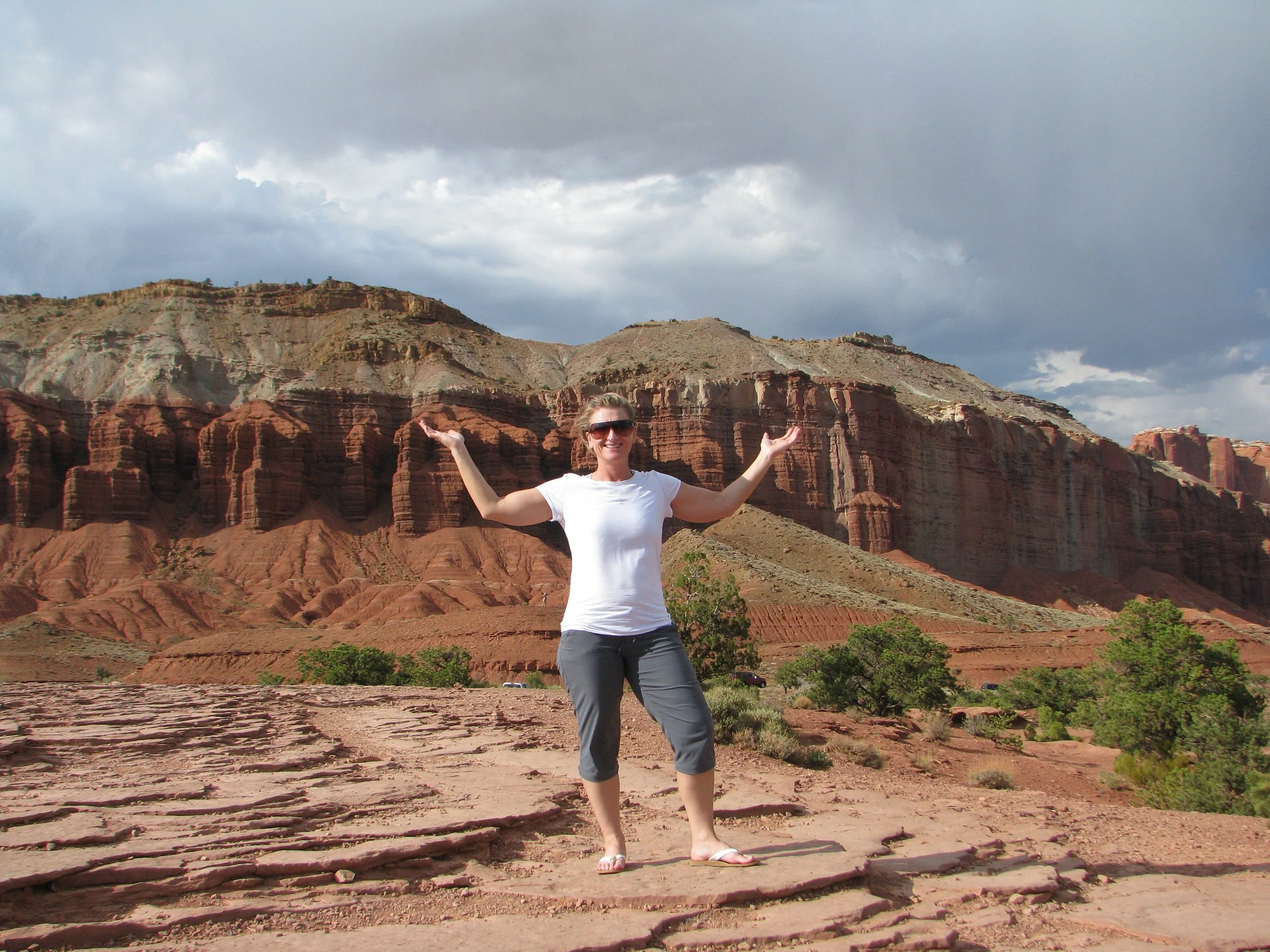 Woman in white T-shirt and gray capri pants standing on rocky terrain with red and orange cliffs and dark cloudy sky in background