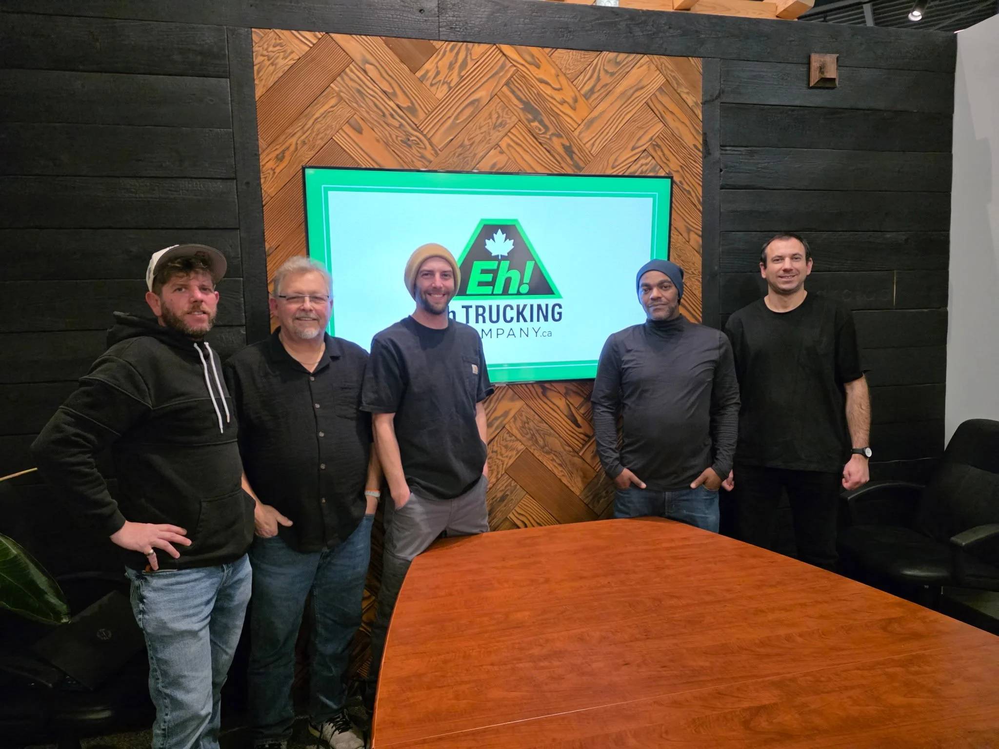 Five men standing in a conference room in front of a digital screen displaying a logo with a maple leaf and the words Eh! Trucking Company.