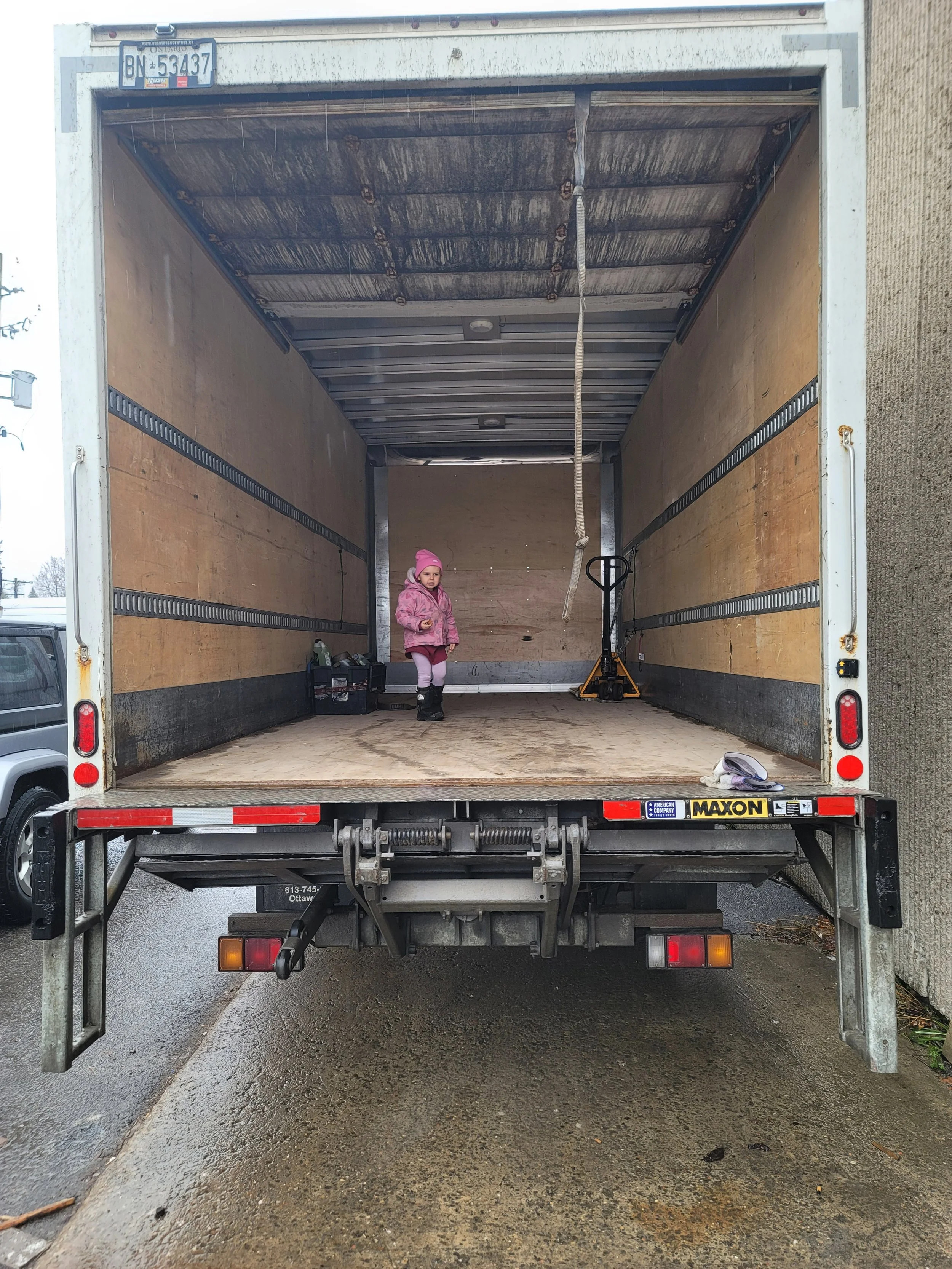 Empty moving truck with a young girl in pink clothing and hat standing inside, with tools and a bottle on the side.