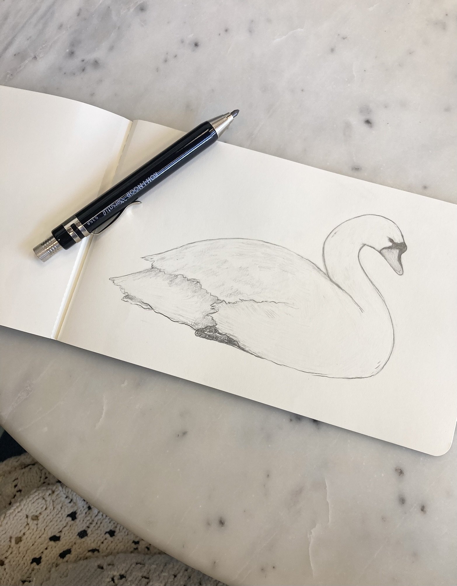 Sketch of a swan on a sheet of paper placed on a marble table, with a black pen resting on the paper.
